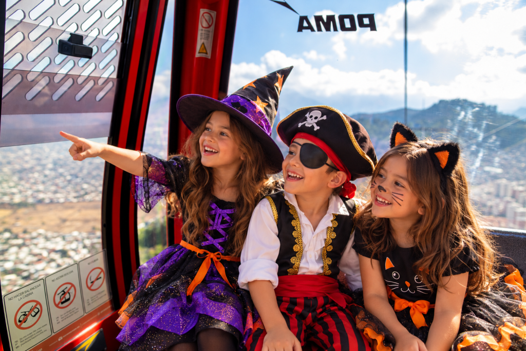 Tres niños sonrientes disfrazados de bruja, pirata y gato negro miran por la ventana de la cabina roja del Teleférico Santiago con vista a la ciudad