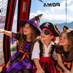 Tres niños sonrientes disfrazados de bruja, pirata y gato negro miran por la ventana de la cabina roja del Teleférico Santiago con vista a la ciudad